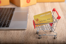 Close-up Shot Of Mock-up Credit Card In Shopping Trolley With Blurred Background Of Laptop And Packaged Boxes On Wooden Table With Copy Space For Ecommerce Business Concept In The Digital Technology.
