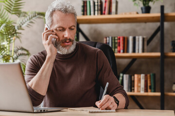 Wall Mural - Handsome businessman in casual clothes talking on mobile phone and making notes while sitting in his office