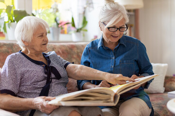 Wall Mural - Senior woman and her adult daughter looking at photo album together on couch in living room
