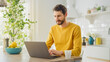 © Gorodenkoff - Young Handsome Man Using Laptop Computer and Drinking Morning Cup of Coffee or Tea While Wearing a Yellow Pullover. Curly-hared Male Sitting in a Modern Sunny Kitchen. Freelancer Working from Home.