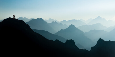 Aufkleber - Man reaching summit after climbing and hiking enjoying freedom and looking towards mountains silhouettes panorama during sunrise.