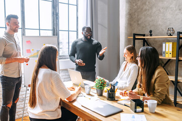 A friendly cheerful atmosphere at the business meeting of a multiracial young team. A young afro-american guy tells something to his colleagues on the meeting at the contemporary office