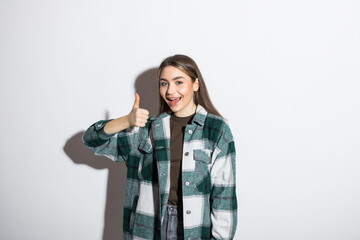 Wall Mural - Portrait of a happy casual girl in plaid shirt standing and showing thumbs up gesture over white background