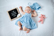 Eleven months old baby girl wearing blue dress laying down on white background with letter board and teddy bear.