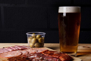 A cold glass of craft beer with a portion of snacks of cold cuts with olives, parma ham and salamis on wooden table over black background. Happy hour time.