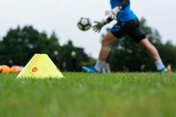 Closeup of yellow marker cone on a grass pitch with a male athlete wearing blue playing football in the background. 