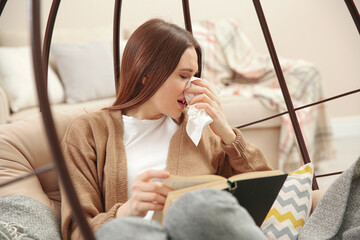 Poster - Sick young woman sneezing while reading book at home. Influenza virus