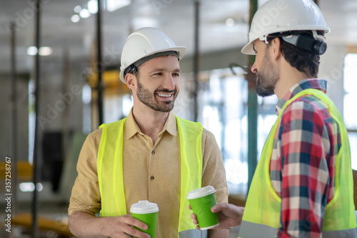 Fotografía Building workers in yellow vests and helmets having coffee break, talking, smili
