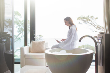 Wall Mural - Woman wearing white bathrobe sitting on edge of modern bathtub using digital tablet