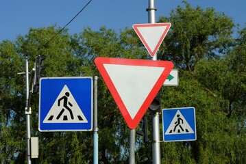 four road signs pedestrian crossing and give way on the street against a background of green branches and a blue sky