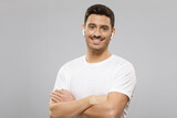 Fototapeta Do przedpokoju - Portrait of young handsome guy in white t-shirt and wireless earphones holding arms crossed, feeling confident at work, isolated on gray background