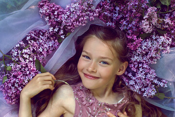 child girl in a purple floral dress lies on the ground among lilacs