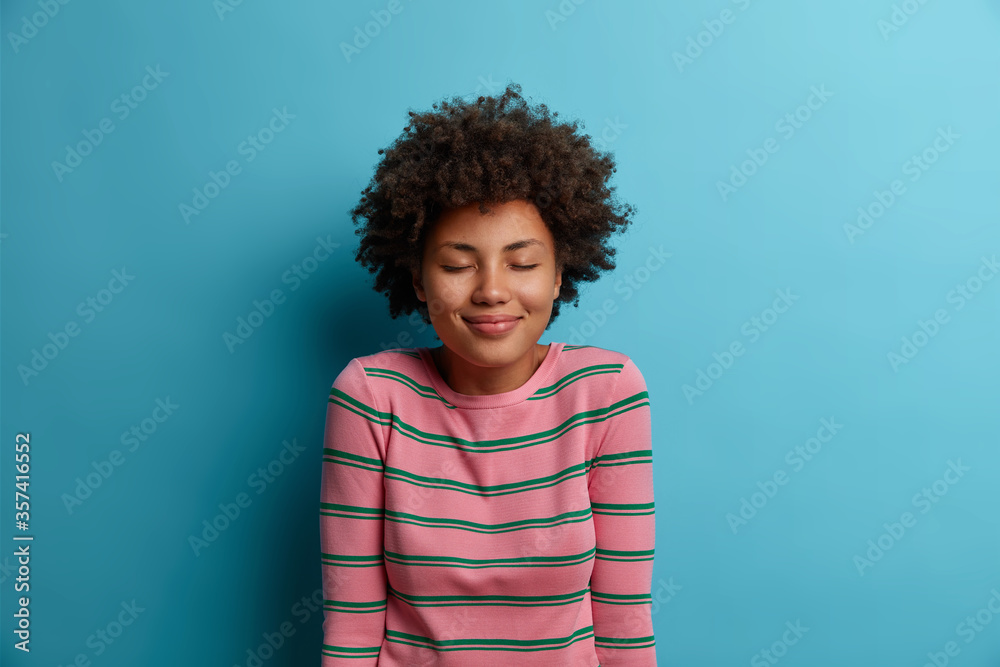 Portrait of pleasant looking dark skinned woman stands with eyes shut, being in good mood, imagines something nice, dreams about perfect vacation, wears striped sweater, isolated on blue background - obrazy, fototapety, plakaty 