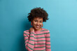 Shy pleased Afro American woman keeps hand on neck, smiles gently, dressed casually, expresses positive emotions, talks casually with someone, isolated on blue background, has optimistic attitude