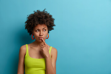 Canvas Print - Serious looking dark skinned woman touches lips and looks thoughtfully aside, has troublesome thoughts, wears casual summer vest, needs think up plan, isolated on blue background, empty space