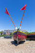 Bow View of Brightly Coloured Fishing Boat on a Stony Beach. Two Poles with Red Flags. Buildings in the Background.  Blue Sky.