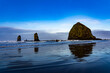 Haystack Rock and the Needles reflected in the wet sand, with blue sky and clouds, Canon Beach, Oregon