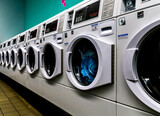 Fototapeta  - Laundromat line of white washing machines with laundry in them and is empty with teal wall and tile floor.