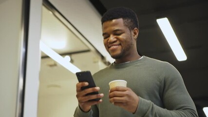 From below medium shot of cheerful young Black man with disposable coffee cup walking down office corridor and enjoying text messaging on cell phone
