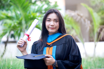 Portrait of a happy and excited young Asian female university graduate wears graduation gown and hat celebrates with a degree in the university campus on the commencement day. Education concept.