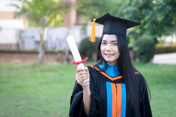 Wall Mural - Portrait of a happy and excited young Asian female university graduate wears graduation gown and hat celebrates with a degree in the university campus on the commencement day. Education concept.