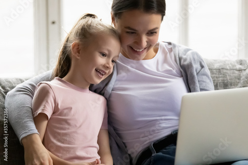 Close up happy mother and little daughter using laptop together, hugging, sitting on cozy couch at home, making video call to relatives, browsing apps, watching cartoons, shopping online