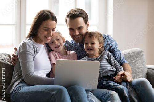 Happy young family with adorable little son and daughter having fun with laptop, looking at screen, sitting on cozy couch at home, watching funny video, playing game, using computer apps together