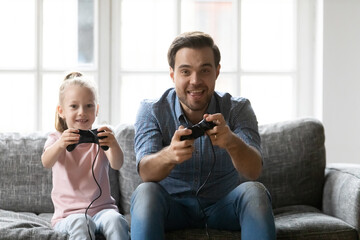 Canvas Print - Happy father and adorable daughter playing video game, holding gamepads controllers, sitting on cozy couch in living room at home, smiling dad and little girl having fun with console