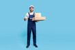© khosrork - Full length cheerful delivery man in blue uniform with gloves pointing cardboard boxes and smiling to camera. Profession of courier, shipment and cargo transportation service. studio shot isolated
