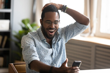 African man sitting at workplace holding smartphone open mouth feels excited by received opportunity, getting hired, salary growth or loan approved read sms from bank, unbelievable news online concept