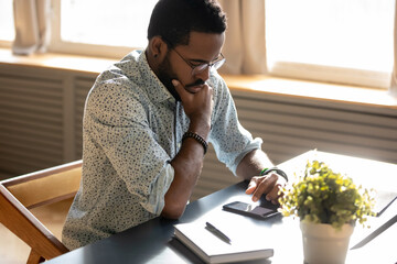 Serious African man read message on smart phone he looks focused or worried by received news, businessman sitting at workplace desk use cellphone websurfing or learn new e-business application concept