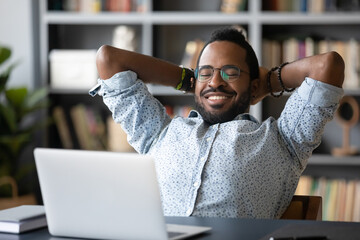 African relaxed guy sit at desk leans on office chair put hands behind head takes break at workplace enjoy videos on notebook. Spend weekend lazy day at home watching movie online streaming service