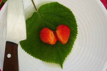 Red strawberry in the shape of a heart, cut in half with a knife, on a green wooden leaf