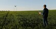 © VAKSMANV - Rear of Caucasian man farmer in hat standing in green wheat field and controlling of drone which flying above margin. Back view on male using tablet device as controller. Technologies in agriculture.