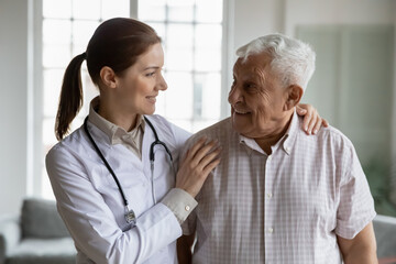 Head shot portrait smiling caring female doctor hugging older man, standing together, young woman caregiver wearing white coat and stethoscope supporting mature senior patient, healthcare concept