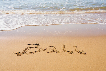 Bali inscription on a tropical sandy beach with waves and foam on a background.