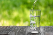 Water is poured into a glass against a background of greenery. A glass of water on a hot summer day with space to copy.