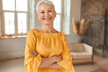 Attractive Confident Mature Blonde Woman Wearing Yellow Dress Crossing Arms On Chest And Smiling Happily At Camera, Posing In Her Newly Renovated Apartment With Sofa And Windows In Background