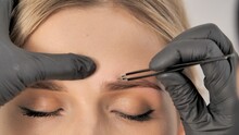 Stylist's Hands In Black Gloves Plucking Eyebrows With Tweezers. Beautiful Attractive Female Face Of A Blonde Well-groomed Woman Or Lady. Styling And Lamination Of Eyebrows. 
