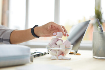 Wall Mural - Close up of young woman enjoying sweet snacks while working at home office, focus on hand holding marshmallow, copy space