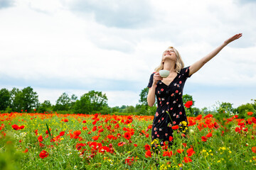 Blonde girl in beautiful dress with cup of coffee in poppies field in summer time