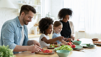 Wall Mural - Full multinational family with cute daughters preparing dietary meal natural nutrition, cutting fresh vegetable for salad, parents caring for children health eat organic food, weekend activity at home