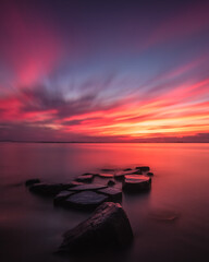 A beautiful sunset lights up the sky as calm smooth water surrounds rocks on the shore. Fire Island New York