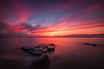 A beautiful sunset lights up the sky as calm smooth water surrounds rocks on the shore. Fire Island New York