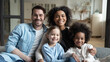 © fizkes - Multinational family married couple and daughters sit on couch looks at camera photo shooting feels happy. Cute multi-ethnic girls and parents portrait, homeowners, new home, prosperous future concept