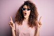 Young beautiful woman with curly hair and piercing wearing funny thug life sunglasses amazed and surprised looking up and pointing with fingers and raised arms.
