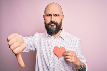 Handsome romantic bald man with beard holding red heart paper over pink background with angry face, negative sign showing dislike with thumbs down, rejection concept