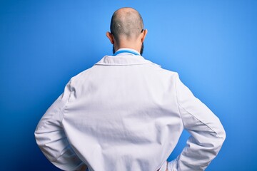 Wall Mural - Handsome bald doctor man with beard wearing glasses and stethoscope over blue background standing backwards looking away with arms on body