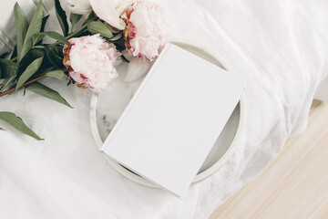 Wall Mural - Wedding still life scene. White empty book cover mockup on marble tray. Pink peony flowers on white linen table cloth. Vintage feminine styled photo, wooden floor. Flat lay. Blurred background.