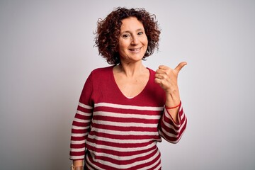 Poster - Middle age beautiful curly hair woman wearing casual striped sweater over white background smiling with happy face looking and pointing to the side with thumb up.